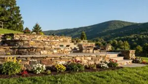 A beautifully crafted retaining wall made of natural stone, seamlessly integrated into a lush East Tennessee landscape. In the foreground, vibrant green grass and colorful blooming flowers complement the wall's earthy tones. The middle ground showcases the sturdy retaining wall, designed with elegant curves and terraces, creating an inviting space for a patio adorned with tasteful outdoor furniture. The background reveals rolling hills and a clear blue sky, typical of the East Tennessee region, bathed in warm afternoon sunlight that casts gentle shadows, enhancing the texture of the stone. The overall mood is serene and inviting, perfect for outdoor relaxation and enjoyment. - Geo Retaining wall contractor